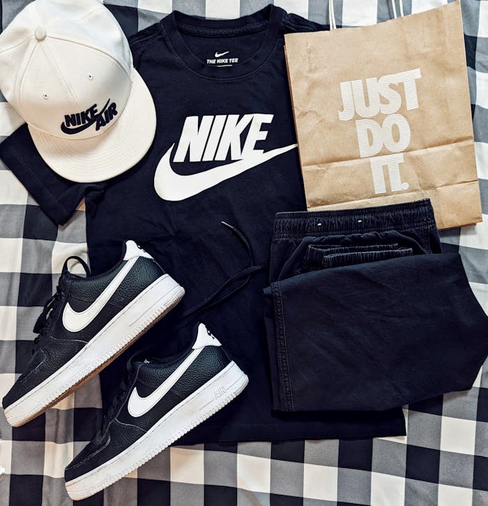 Flat lay of Nike apparel including t-shirt, cap, sneakers, and paper bag on checkered surface.