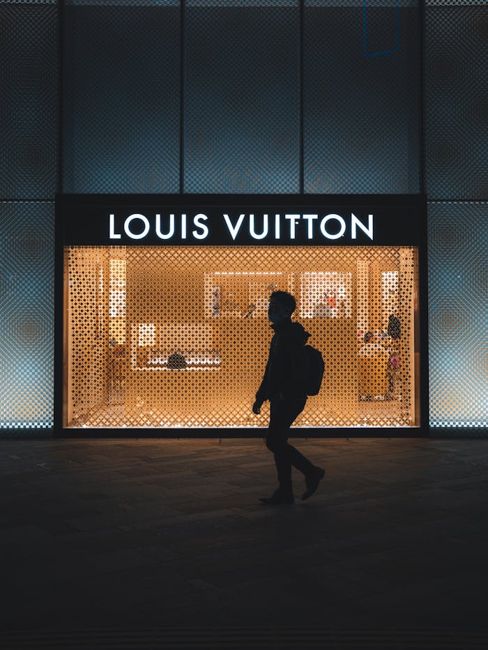 Silhouette of person walking by an illuminated Louis Vuitton storefront at night.